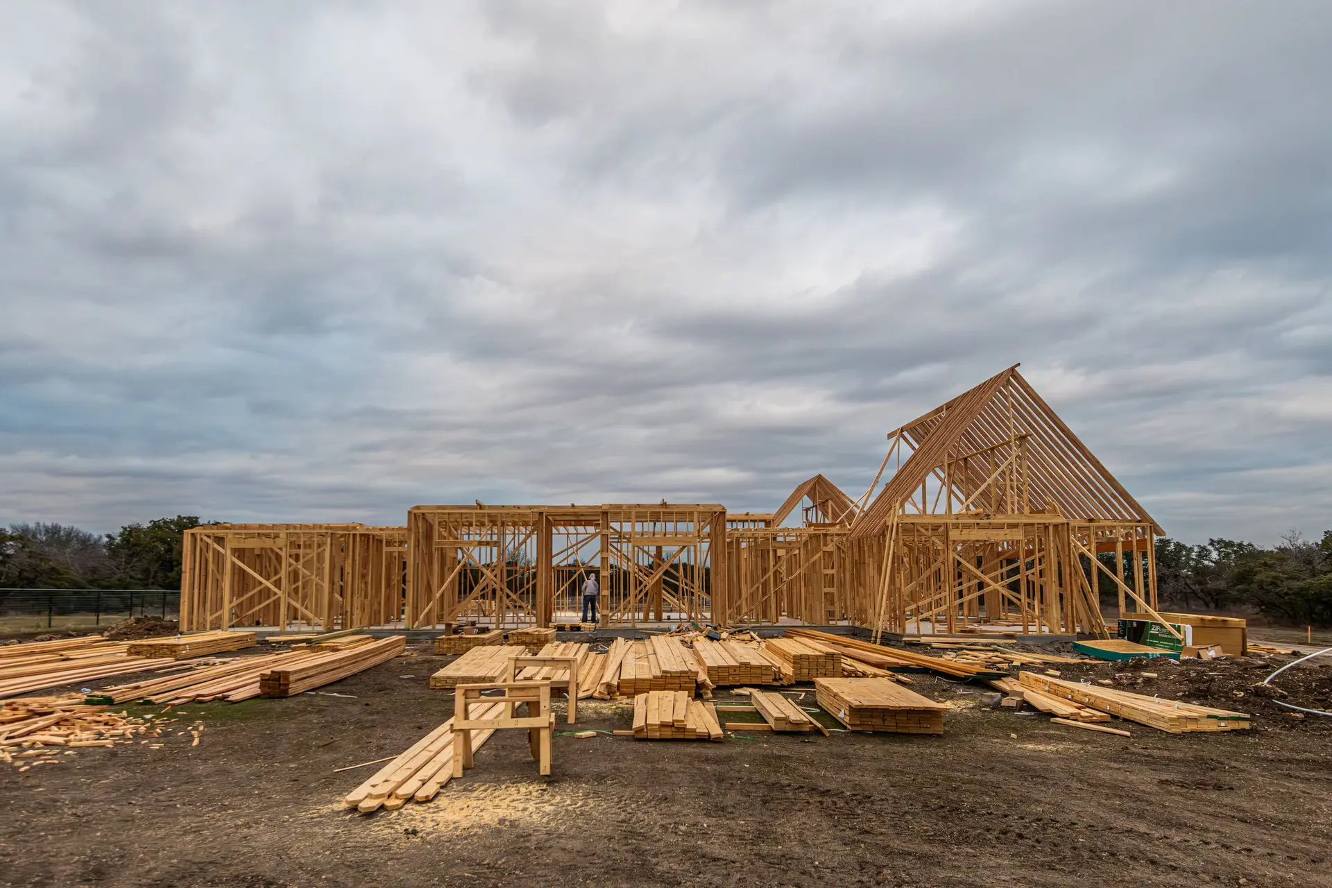 New house framing on a lot stacked with lumber under a gray sky